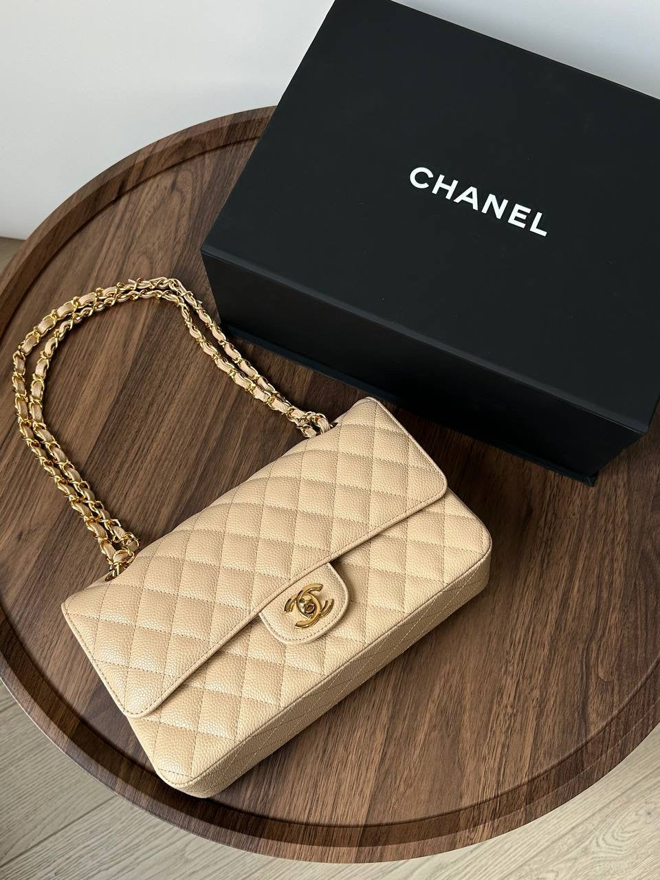 Beige quilted handbag with gold chain and Chanel box on a wooden surface