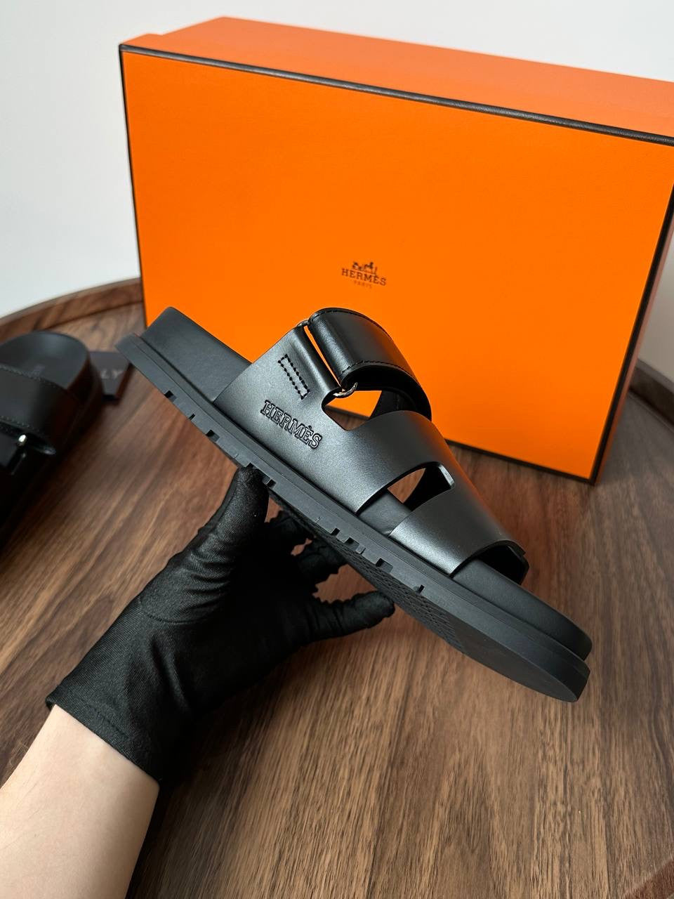 Black sandal held by a gloved hand with an orange Hermès box in the background.
