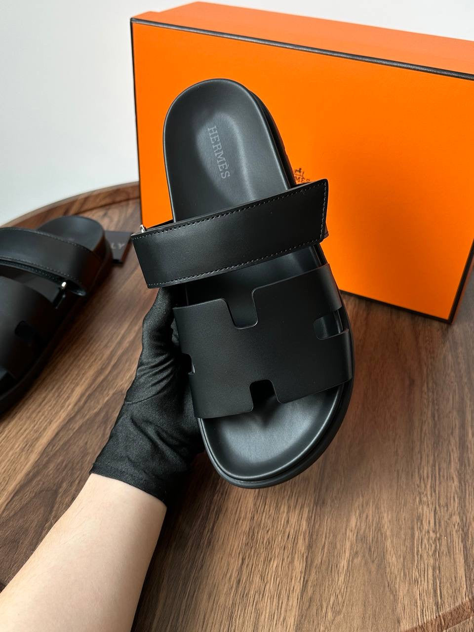 Black sandal held by a gloved hand with an orange Hermes box in the background.